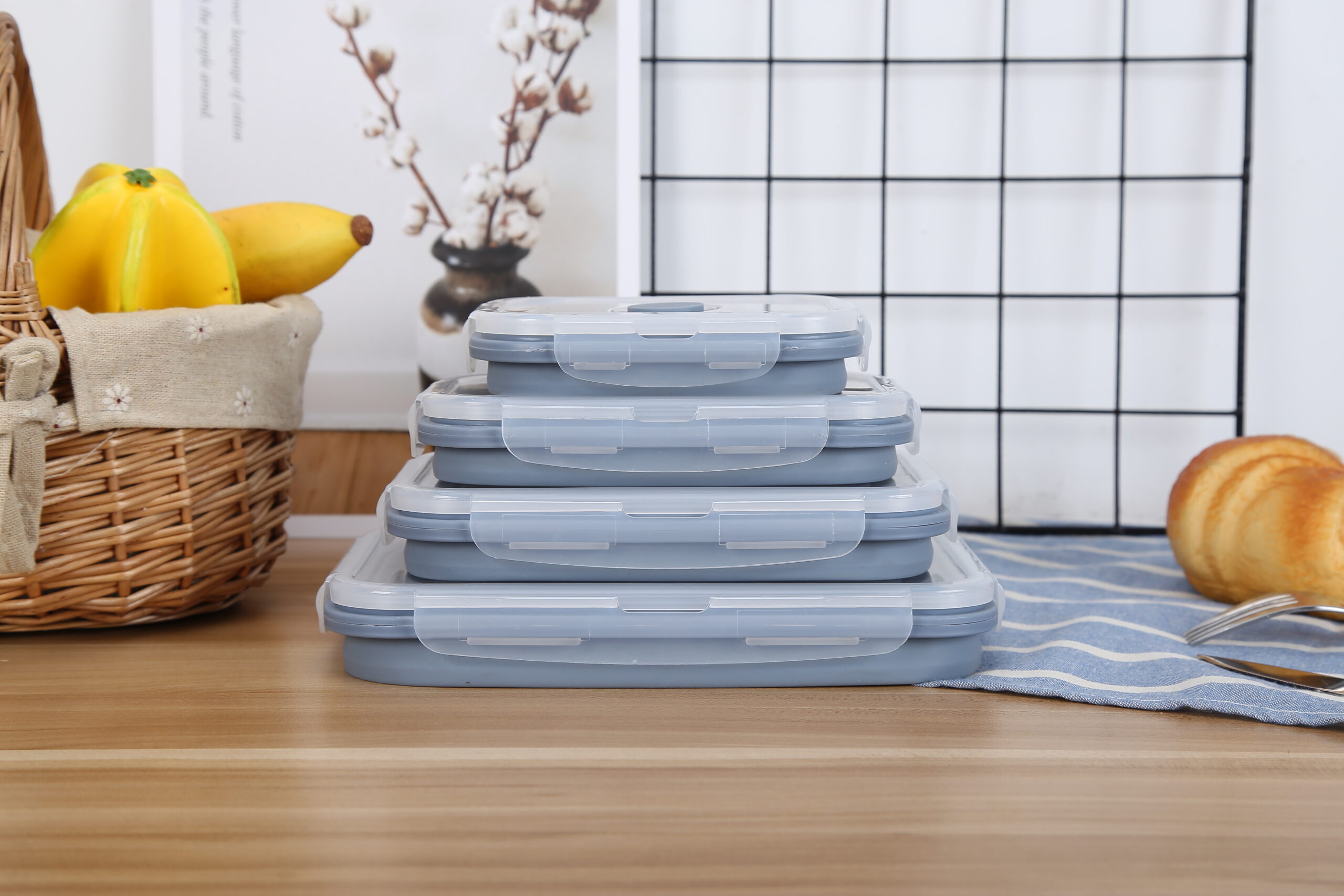 Set Of 4 Collapsible Food Containers (Pre Order Listing – Dispatch Date 1St May) On A Wooden Countertop With A Basket Of Bananas And A Croissant Placed Nearby, Against A Backdrop Of A White Grid Screen.